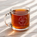 A glass mug filled with a warm, amber-colored liquid, likely tea, sits on a wooden surface. The mug has a message engraved on it that reads "Happy Mother's Day I love you!"