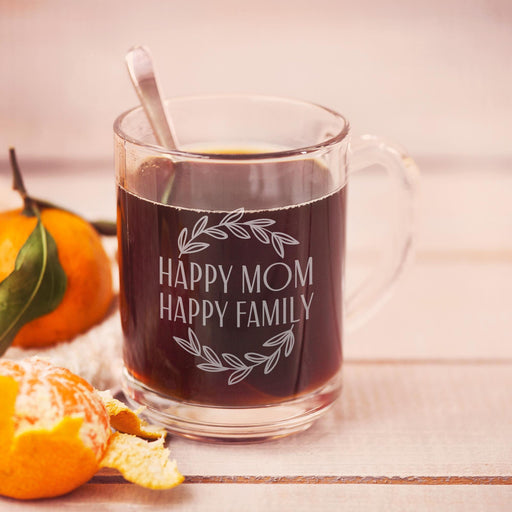 A glass mug filled with a dark liquid, likely coffee, sits on a wooden surface. The mug has a white floral design and the text "HAPPY MOM HAPPY FAMILY" engraved on it.