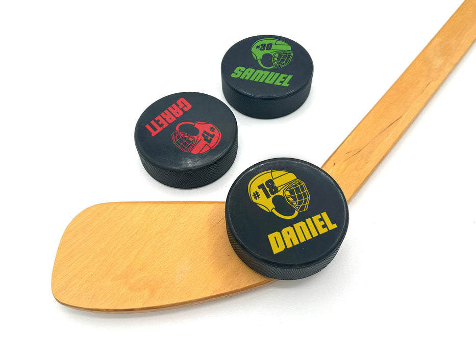 three hockey pucks with name & number helmet designs printed on them with the colors green, red, & yellow with the names Samuel, Garett, and Daniel