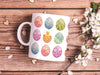 15 oz white ceramic mug with an easter pattern of floral decorated eggs with a baby chick popping out of an egg on a wooden table surrounded by white flowers and heart shaped petals