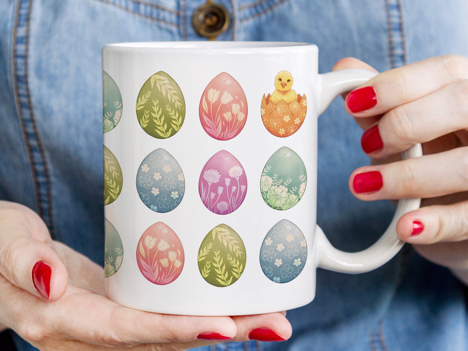 woman with red painted nails holding a 15 oz white ceramic mug with an easter pattern of floral decorated eggs with a baby chick popping out of an egg