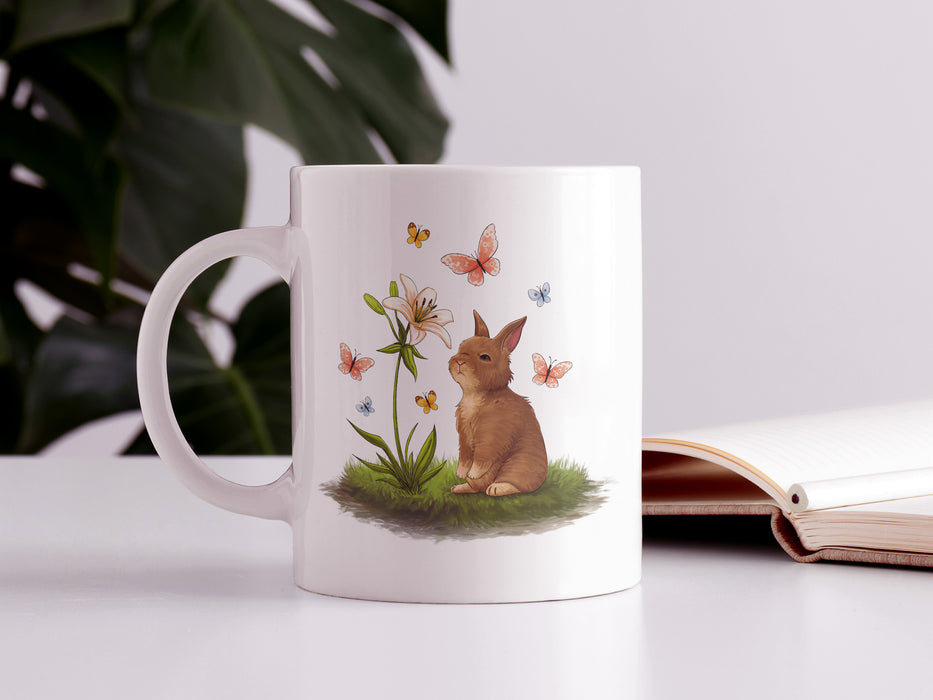 15 ounce white ceramic mug with easter artwork of a baby bunny and a lily surrounded by butterflies on a white table next to an open book and a house plant