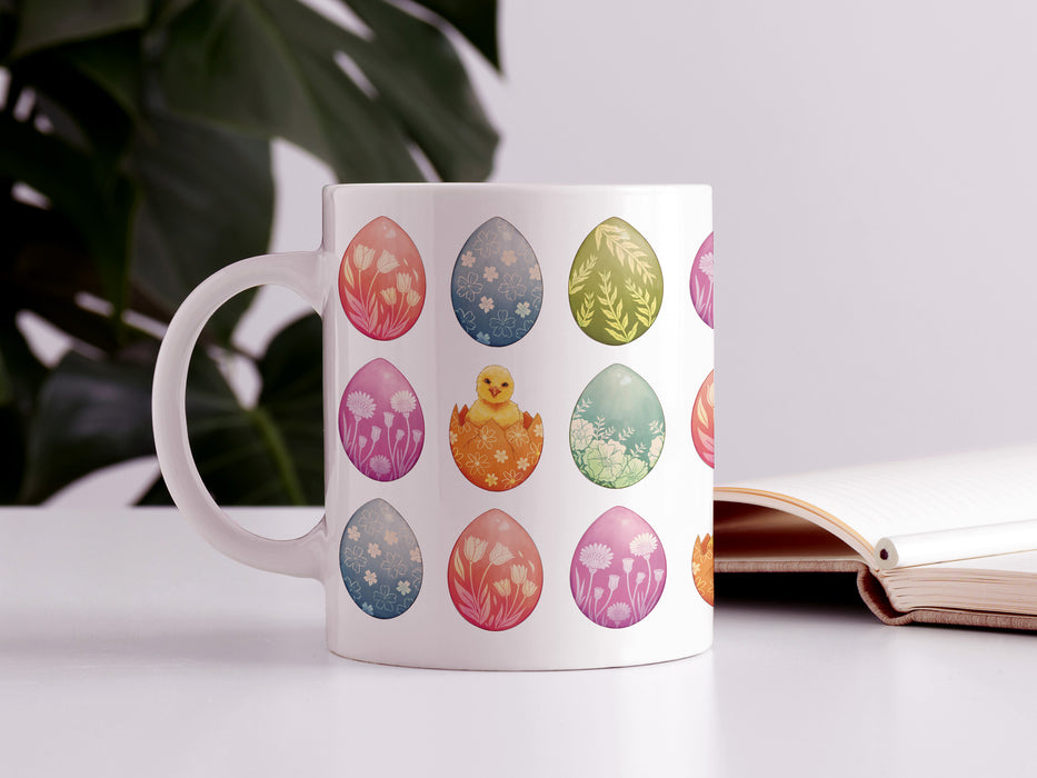 15 oz white ceramic mug with an easter pattern of floral decorated eggs with a baby chick popping out of an egg on a white table next too an open book and a house plant
