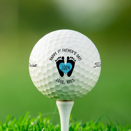Single white Titleist golf ball with first Father's Day footprint design on white golf tee with golf course grass in the background