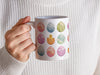 woman in a white sweater holding a 15 oz white ceramic mug with an easter pattern of floral decorated eggs with a baby chick popping out of an egg