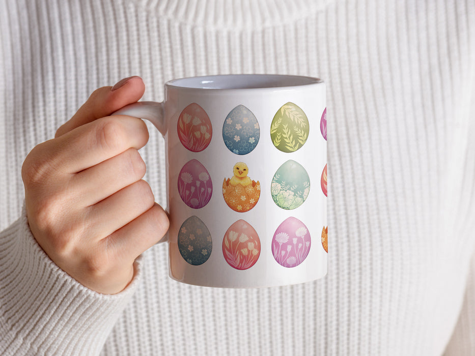 woman in a white sweater holding a 15 oz white ceramic mug with an easter pattern of floral decorated eggs with a baby chick popping out of an egg