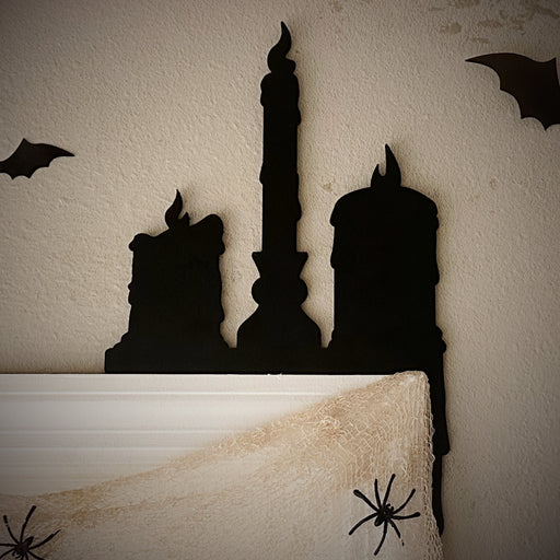 A black door frame topper, designed as a silhouette of three dripping wax candles, is seen on top of a white door frame. Spooky bats and spiders surround the topper. The wall behind is beige.