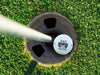 Single white titleist golf ball with Kick Putt Dad design in golf course hole next to pole surrounded by grass