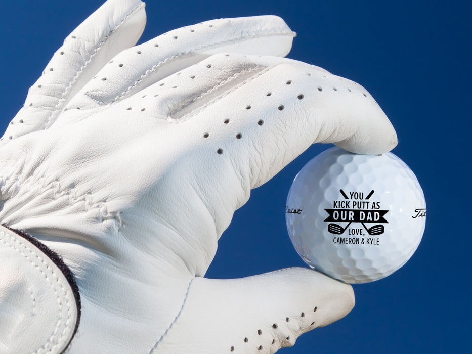 White gloved hand holding single white titleist golf ball with Kick Putt Dad design in front of dark blue background