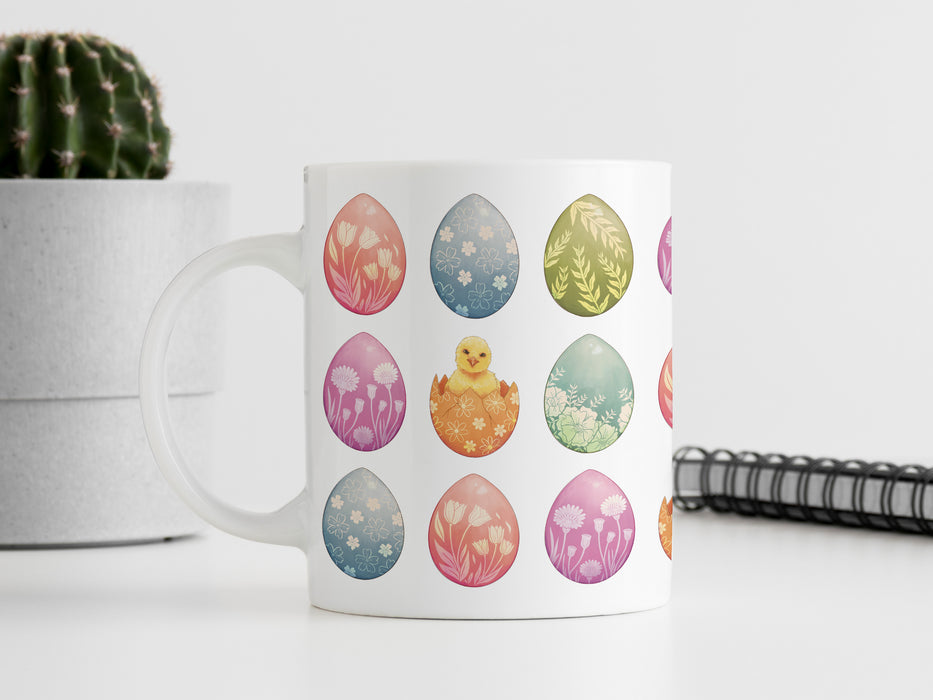 15 oz white ceramic mug with an easter pattern of floral decorated eggs with a baby chick popping out of an egg on a white table next to a spiral notebook and a potted cactus