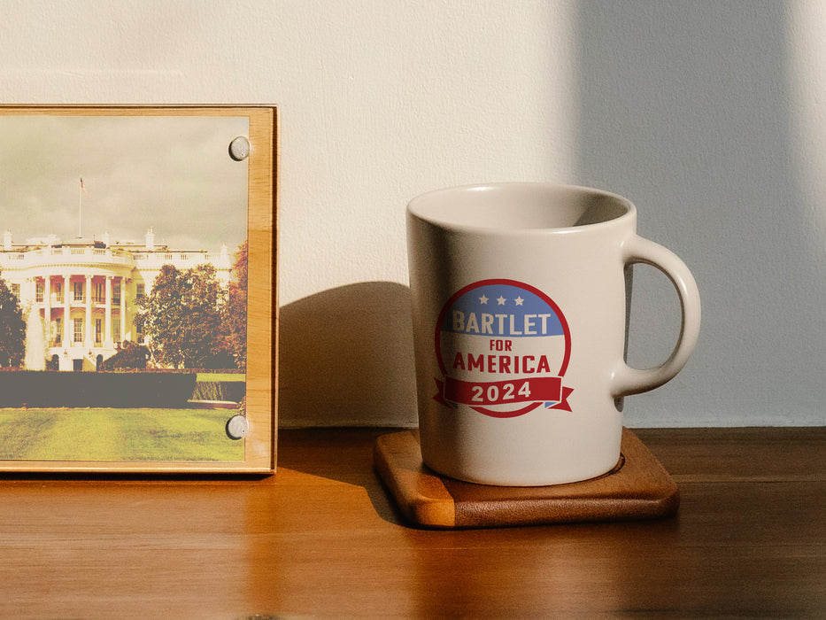 white mug on wooden desk ontop of a wooden coaster next to a wooden frame of the whitehouse mug has red white and blue American flag design with typography that says Bartlet for America 2024