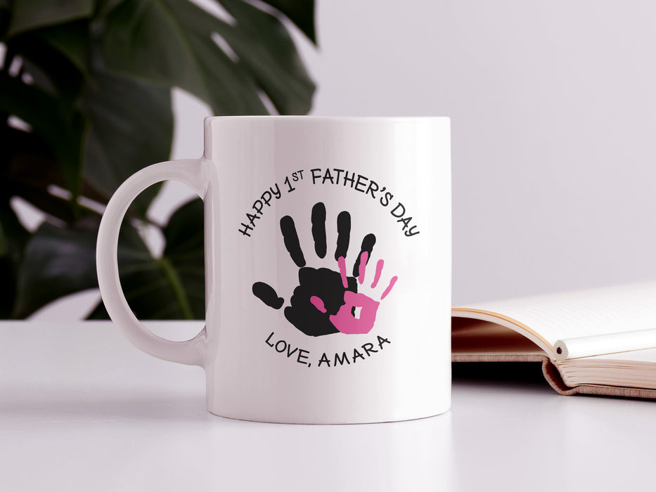 white ceramic mug that says happy first fathers day love Amara with a pink handprint design on a white table next to an open book and a house plant