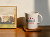 White mug on wooden coaster ontop of wooden table next to wooden photo frame with a picture of the White House  mug has red white and blue American design with red stars that has typography that says Bartlet for America