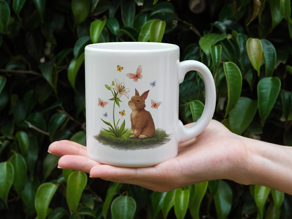 hand holding a 15 ounce white ceramic mug with easter artwork of a baby bunny and a lily surrounded by butterflies in front of outdoor plants