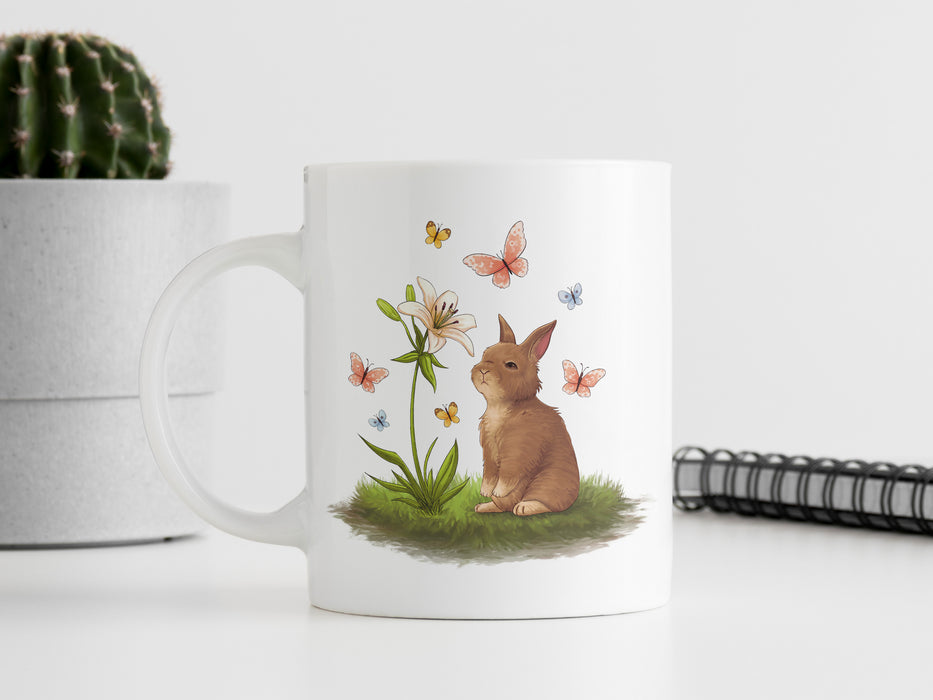 15 ounce white ceramic mug with easter artwork of a baby bunny and a lily surrounded by butterflies on a white table next to a spiral note book and a potted cactus