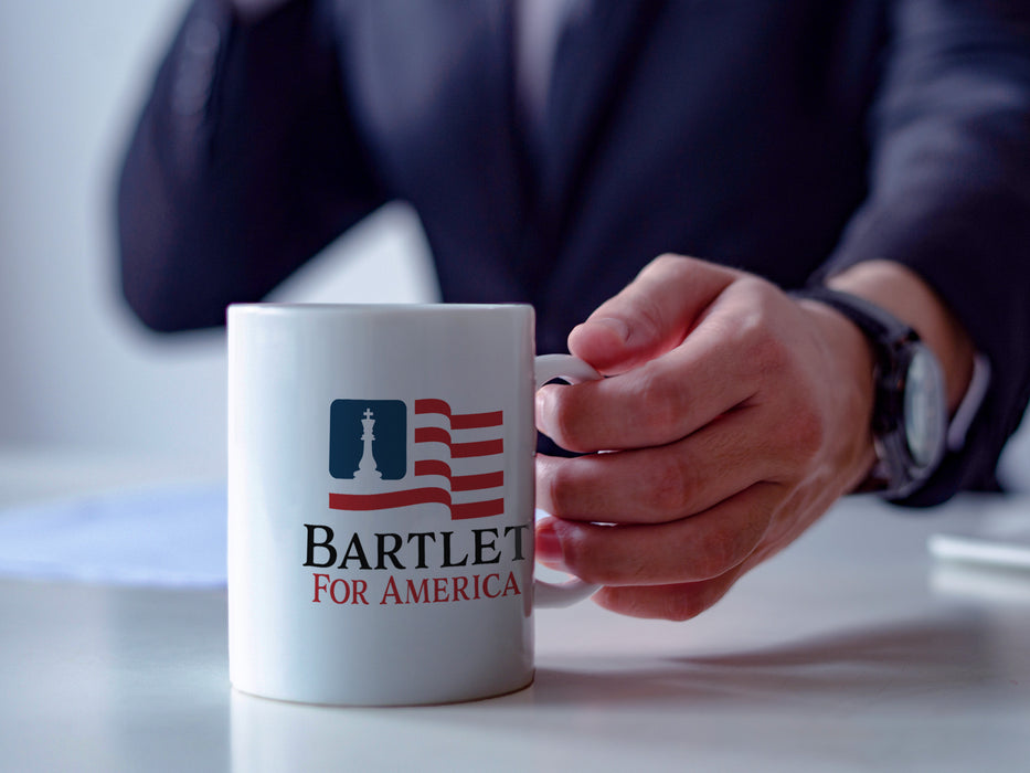 business man sitting at white desk holding a white mug that has red white and blue patriotic American flag design with a king chess piece with Typography that says Bartlet for America