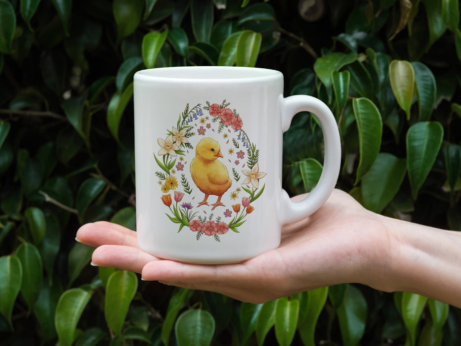 Hand holding 15 ounce white ceramic mug with spring easter art of a baby chick surrounded by various colorful flowers in front of outdoor plants