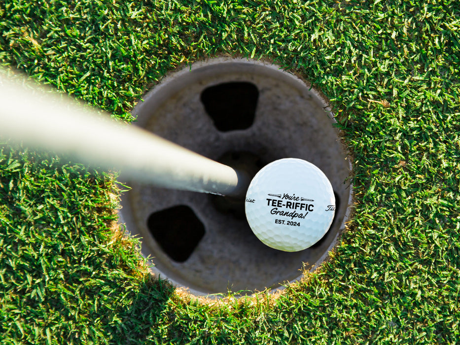 Single white titleist golf ball with Tee-riffic Grandpa design in golf course hole next to pole surrounded by grass