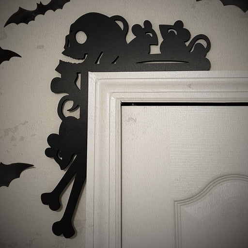 A black door frame topper, designed with the silhouette of scurrying rats, bones, and a skull, is seen on top of a white door frame. The beige wall behind has fake bats attached to it.