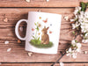 15 ounce white ceramic mug with easter artwork of a baby bunny and a lily surrounded by butterflies on a wooden table next to a bunch of little flowers and heart shaped petals