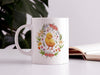 15 ounce white ceramic mug with spring easter art of a baby chick surrounded by various colorful flowers on a white table next to a house plant and an open book