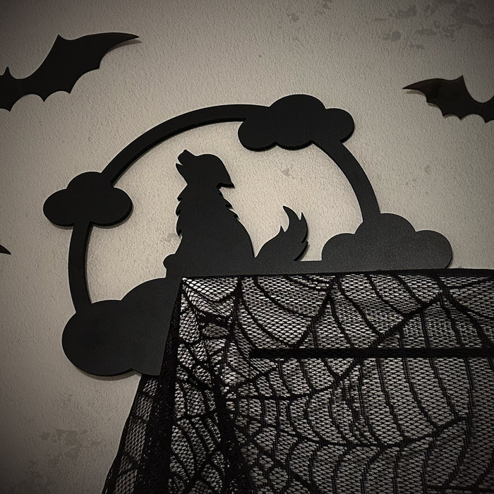A black door frame topper, designed with the silhouette of a howling wolf, full moon, and clouds, is seen on top of a white door frame. The frame is covered in black spider webs and fake spiders. The beige wall behind has fake bats attached to it.