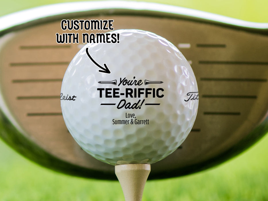 Single white titleist golf ball with Tee-riffic Dad design on wooden golf tee with golf club and golf course grass in the background. The text "customize with names" is above the ball with an arrow pointing toward it.