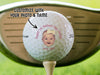 Single white titleist golf ball with first Father's Day baby photo design on wooden golf tee with golf club and golf course grass in the background. The text "customize with your photo and name" is above the ball with an arrow pointing toward it.