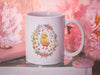 15 ounce white ceramic mug with spring easter art of a baby chick surrounded by various colorful flowers sitting on a book surrounded by various pink items such as a wallet, wallpaper, and a pink flower