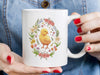 woman with red painted nail polish holding a 15 ounce white ceramic mug with spring easter art of a baby chick surrounded by various colorful flowers