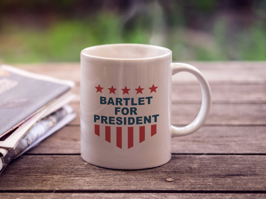 white mug with red white and blue American design with typography that says Bartlet for president with Stars and Stripes on a wooden table outside next to a newspaper with hot coffee steam