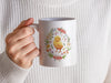 woman in a white sweater holding a 15 ounce white ceramic mug with spring easter art of a baby chick surrounded by various colorful flowers
