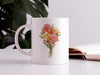 15 ounce ceramic mug featuring artwork of a bouquet of pastel spring flowers on a white table next to an open book and a house plant