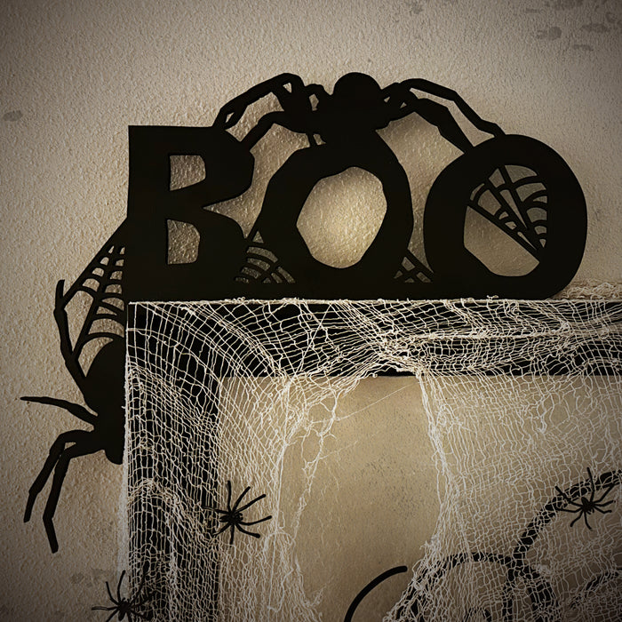 A black door frame topper, designed with the word BOO, spiders, and spider webs, is seen on top of black frame. Spooky spiders and spider webs are attached to the frame. The wall behind is beige.