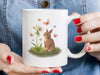 woman with red painted nails holding a 15 ounce white ceramic mug with easter artwork of a baby bunny and a lily surrounded by butterflies