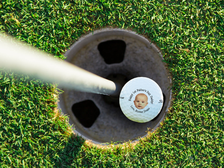 Single white titleist golf ball with first Father's Day baby photo design in golf course hole next to pole surrounded by grass