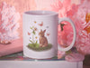 15 ounce white ceramic mug with easter artwork of a baby bunny and a lily surrounded by butterflies sitting ontop of a book next to pink items such as a wallet, a pendant, and a flower