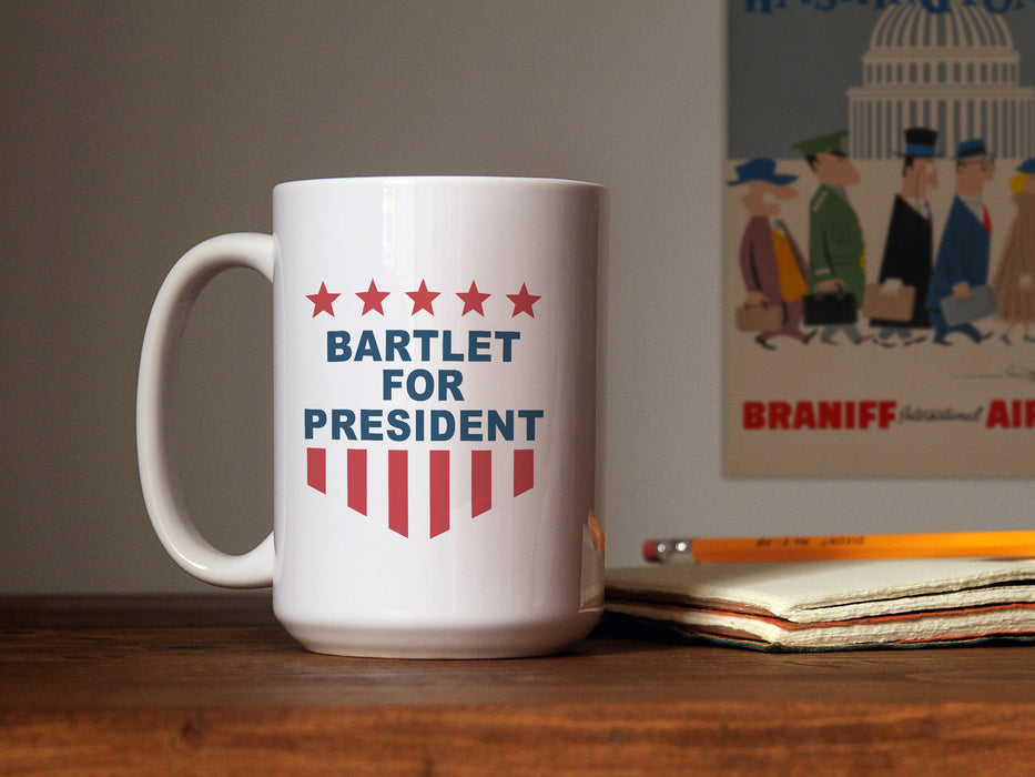 white mug with red white and blue American design with typography that says Bartlet for president with Stars and Stripes ontop of wooden counter next to notebook with wooden pencil in front of white wall with cartoon poster of the capital