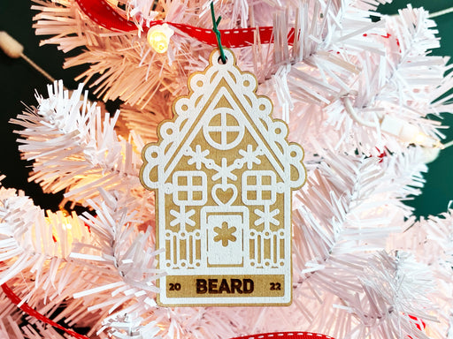Ornament shown hanging from Christmas tree. The ornament is wooden and shaped like a gingerbread house. A name and year are laser engraved into ornament.