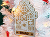 Ornament shown hanging from Christmas tree. The ornament is wooden and shaped like a gingerbread house. A name and year are laser engraved into ornament.