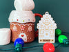 Ornament is shown sitting on a table next to a gingerbread house mug. The ornament is wooden and shaped like a gingerbread house. A name and year are laser engraved into the ornament.