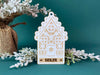 Ornament shown on a table with a Christmas tree and branches. The ornament is wooden and shaped like a gingerbread house. A name and year are laser engraved into ornament.