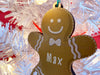 Ornament shown hanging from Christmas tree. The ornament is wooden and shaped like a gingerbread boy. Name and year are written on ornament in white.