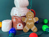 Ornament is shown sitting on a table next to a gingerbread house mug. The ornament is wooden and shaped like a gingerbread boy. Name and year are written on ornament in white.