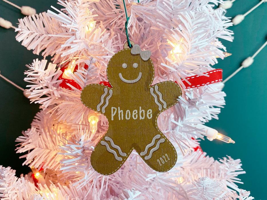 Ornament shown hanging from Christmas tree. The ornament is wooden and shaped like a gingerbread girl. Name and year are written on ornament in white.