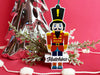  Ornament shown sitting on table next to Christmas tree and branches. The ornament made from PVC and shaped like a nutcracker. A last name and year are written on the ornament.
