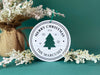 Ornament shown sitting on table next to Christmas tree and branches. The ornament is wooden and shaped like a circle with a Christmas Tree cutout in the middle. Ornament says Merry Christmas, Year, and Family Name