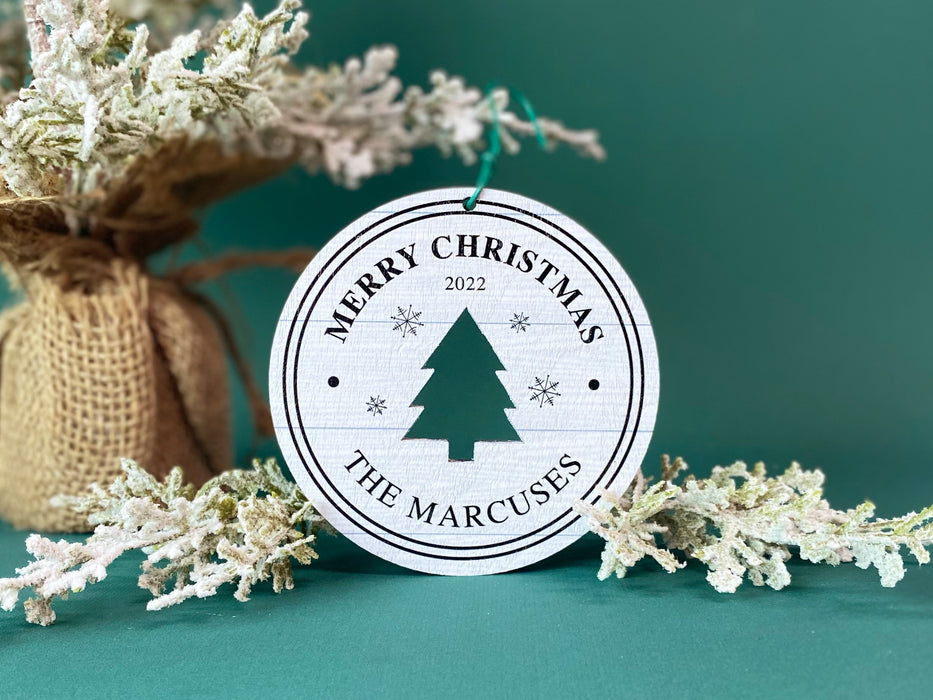 Ornament shown sitting on table next to Christmas tree and branches. The ornament is wooden and shaped like a circle with a Christmas Tree cutout in the middle. Ornament says Merry Christmas, Year, and Family Name
