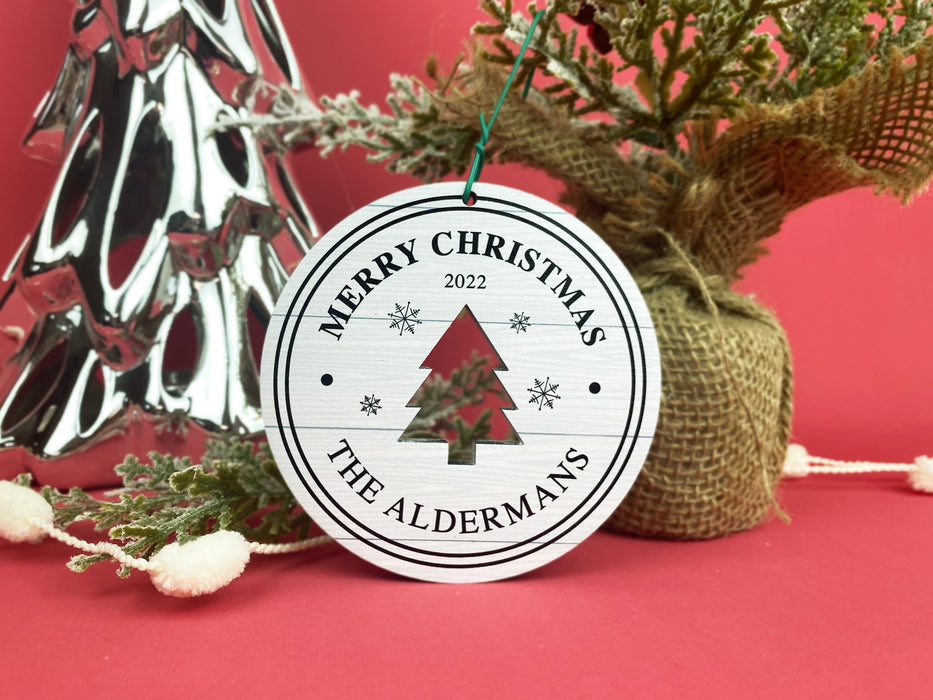 Ornament shown sitting on table next to Christmas trees and branches. The ornament is wooden and shaped like a circle with a Christmas Tree cutout in the middle. Ornament says Merry Christmas, Year, and Family Name