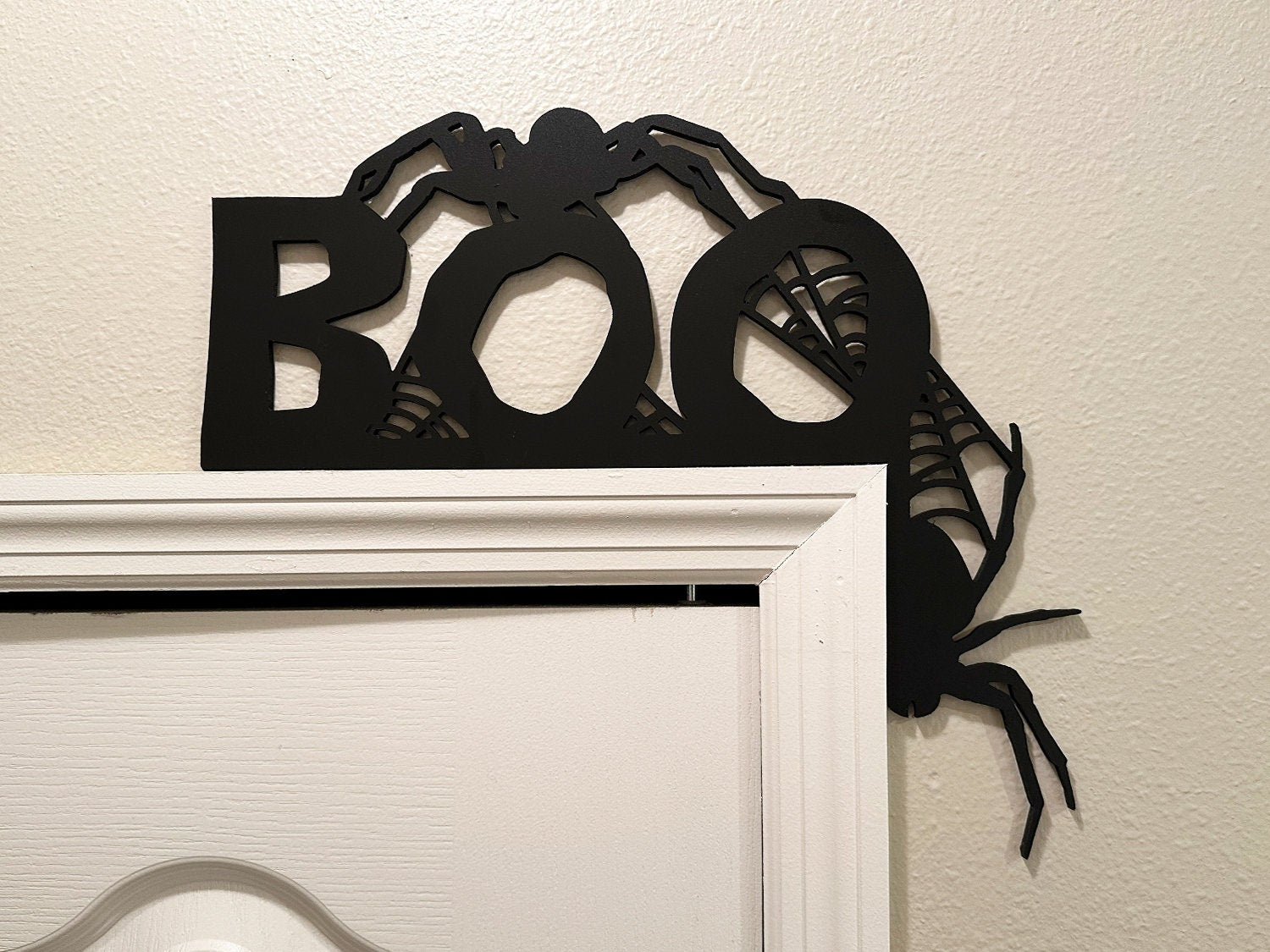 A black door frame topper, designed with the word BOO, spiders, and spider webs, is seen on top of black frame. Spooky spiders and spider webs are attached to the frame. The wall behind is beige.